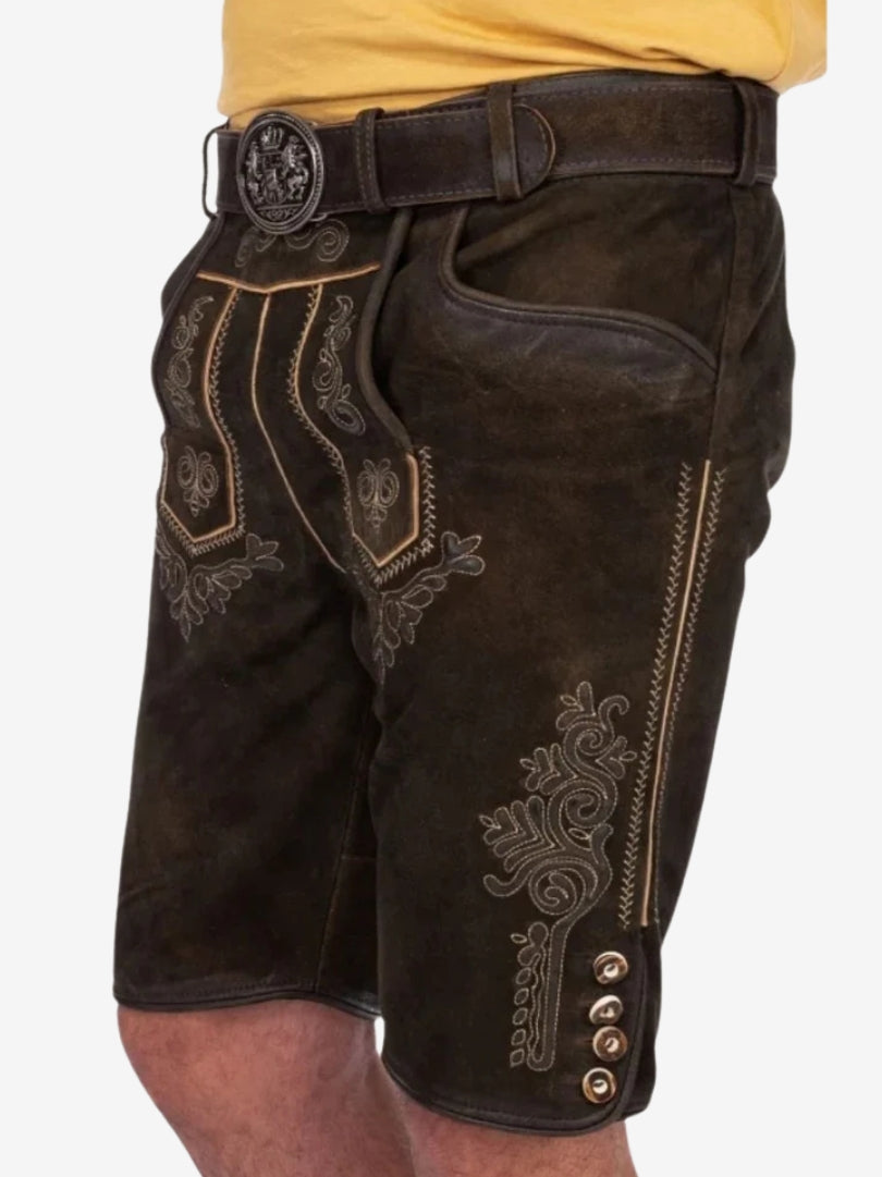 Angled front view of dark brown lederhosen with detailed embroidery, front flap panel, side pocket, and belt with ornate buckle.
