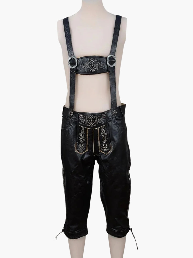Black leather bundhosen with suspenders displayed on a mannequin, featuring embroidered front flap and decorative metal buckles.