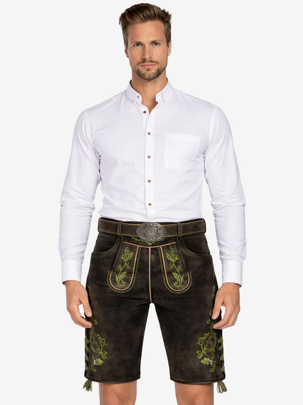 A man wearing traditional brown leather lederhosen with green embroidery, paired with a white button-up shirt, standing against a plain background.