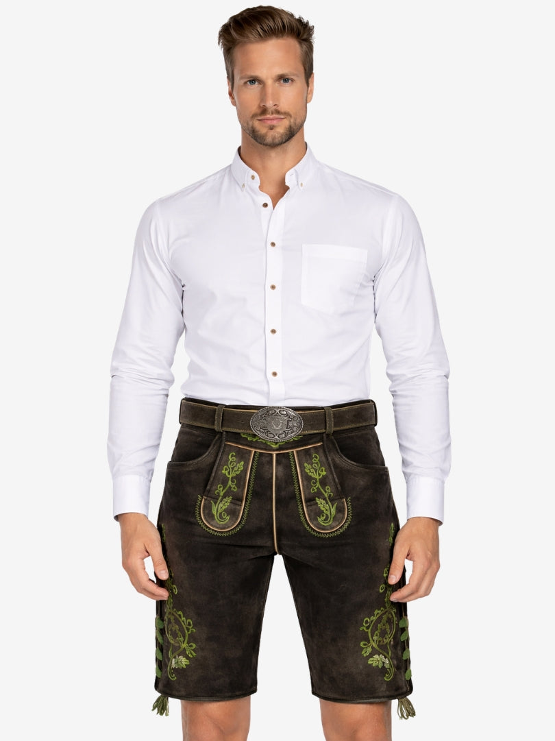 A man wearing traditional brown leather lederhosen with green embroidery, paired with a white button-up shirt, standing against a plain background.