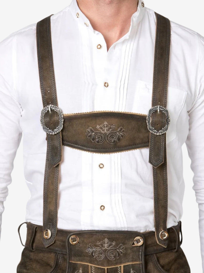 Close-up front view of brown lederhosen suspenders with decorative metal buckles worn over a white shirt.