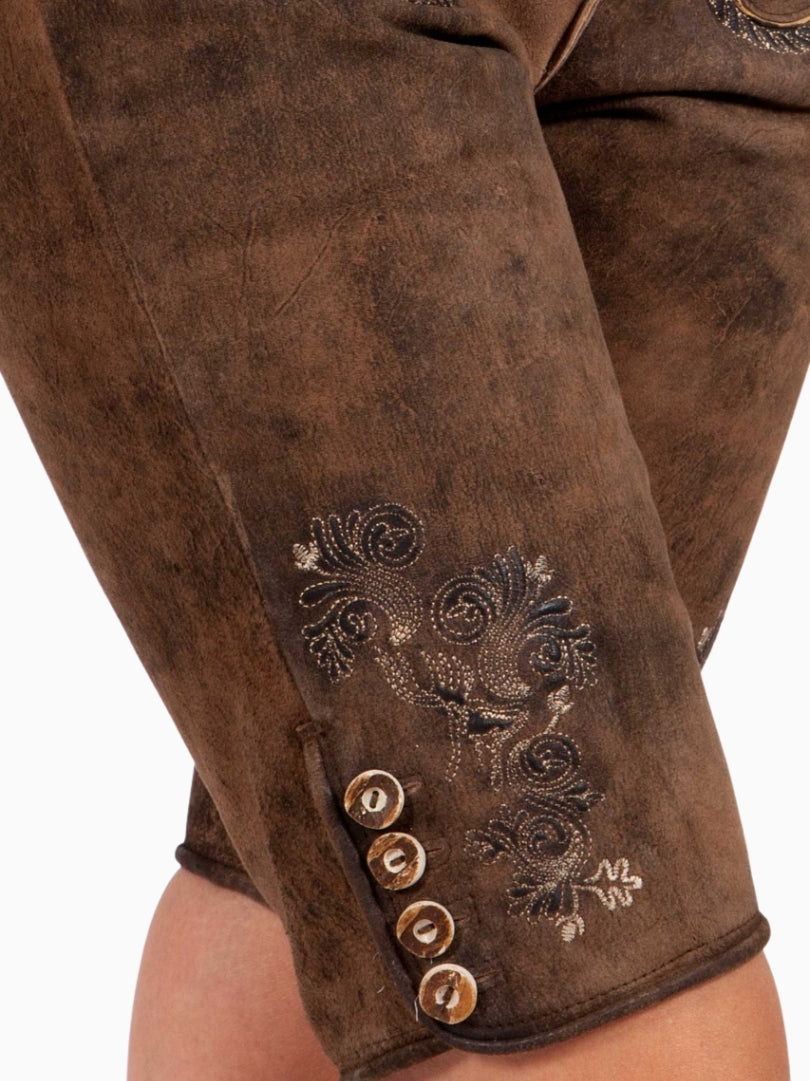 Close-up side view of rustic brown lederhosen-style shorts showing embroidered floral detailing and decorative buttons near the hem on a textured leather finish.