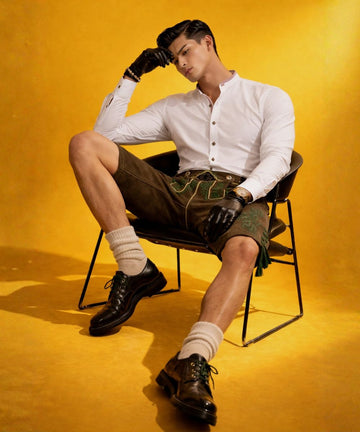 Modern lederhosen outfit for Oktoberfest 2026 featuring a stylish male model wearing a white Bavarian shirt paired with brown embroidered leather lederhosen, sitting confidently on a chair against a vibrant yellow background, showcasing traditional German fashion with a modern twist, perfect Oktoberfest men’s outfit inspiration.