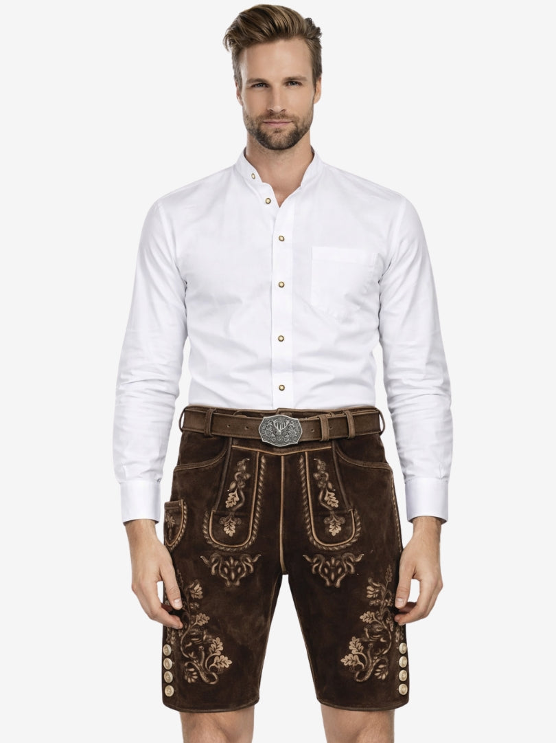 Man wearing a white button-up shirt paired with dark brown embroidered lederhosen and a traditional belt, standing against a plain background.