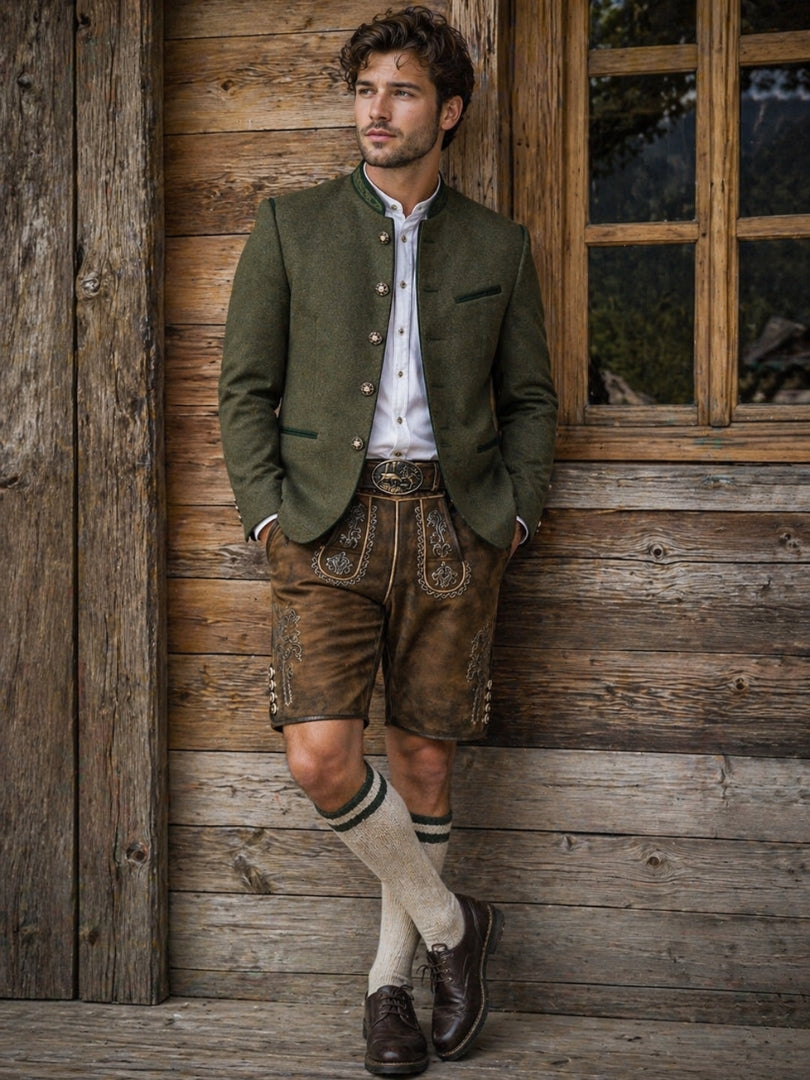 Man wearing traditional Bavarian outfit with green trachten jacket, leather lederhosen shorts, white shirt, knee-high socks, and brown shoes standing against a rustic wooden wall.