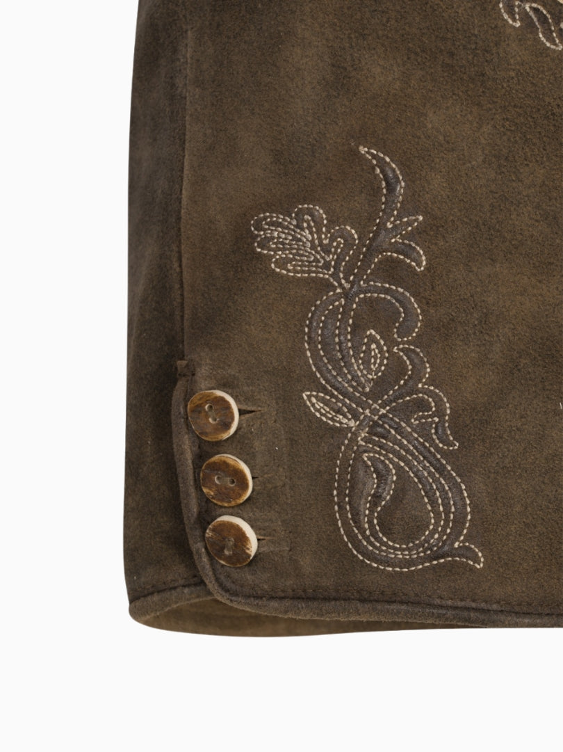 Close-up side view of rustic brown lederhosen-style shorts showing embroidered floral detailing and decorative buttons near the hem on a textured suede finish.
