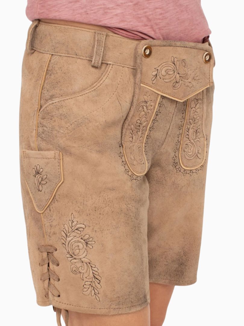Side view of beige traditional alpine lederhosen-style shorts with embroidered detailing, decorative front flap with buttons, side lacing, and a suede-like textured finish, worn with a pink top